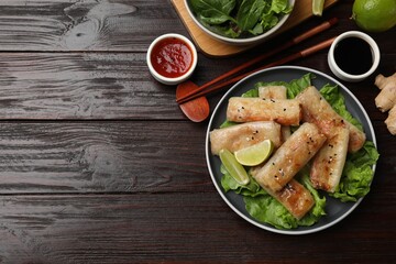 Tasty fried spring rolls, lettuce, sauce and lime on wooden table, flat lay. Space for text