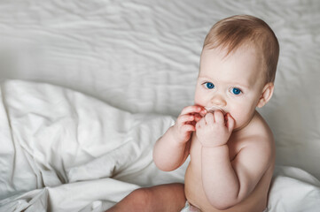 Portrait of beautiful baby 11 months old in diaper on bed close-up. Skincare, healthcare concept for children. Teething, hands on toddler's mouth