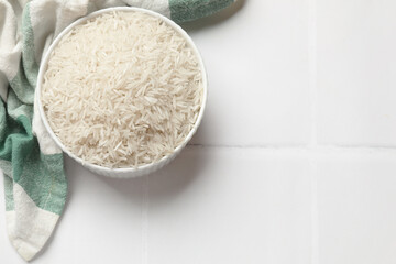 Raw basmati rice in bowl on white tiled table, top view. Space for text