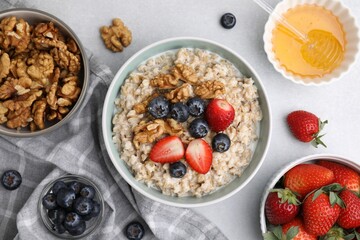 Tasty oatmeal with strawberries, blueberries and walnuts surrounded by ingredients on grey table, flat lay