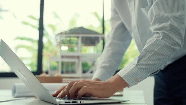 Close up of professional male architect engineer measure house model and using laptop analysis data at meeting table with house model and map . scatter around. Focus on hand. Immaculate.