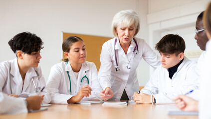 Group of medical workers in white coats sitting around desk during professional training course, listening to experienced aged female lecturer