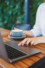 Hands of a woman typing on a laptop keyboard with a cup of coffee, phone, notes on the table. A college student on the terrace of a cafe-restaurant.