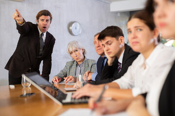 Angry boss scolding upset subordinates sitting at table in office meeting room, expressing dissatisfaction with work