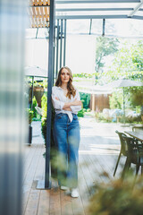 A cute young woman in a white shirt and blue jeans is standing on the terrace of a summer cafe. Woman in casual stylish summer clothes