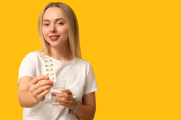 Young woman with vitamin A pills and glass of water on yellow background