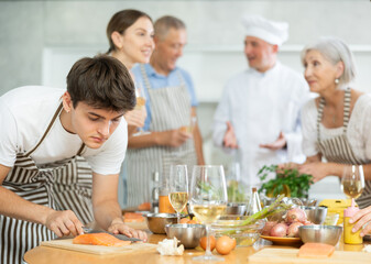 Positive interested young guy attending group culinary classes, standing by table with ingredients and utensils, learning culinary skills from professional chef