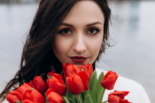 Attractive brunette woman walking on the beach shore in moody cloudy windy weather with bouquet of red tulips flowers, dressed in white suit jacket. International Women`s Day 8th March concept