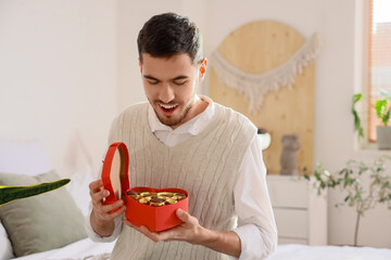 Handsome man with box of heart-shaped chocolate candies in bedroom