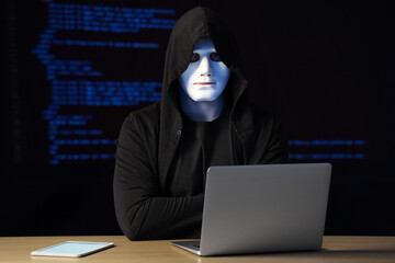 Hacker with laptop at table against programming code on dark background