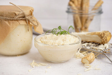 Jar and bowl of horseradish sauce with horseradish roots on white background