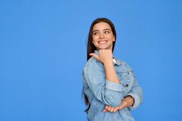 Smiling pretty gen z cute happy 20s Latin teen student girl with brunette hair wearing denim jacket pointing aside presenting ads standing isolated on blue background. Copy space.
