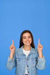 Happy cute Latin teenage student girl pointing up standing on background. Smiling pretty hispanic teenager with brunette hair wearing denim jacket presenting ads standing isolated on blue. Vertical