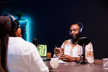Obraz premium African american couple wearing wireless headphones are making a podcast at home using audio gadgets. Black male host interviewing female blogger for his talk show online audience.