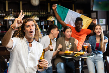 Male Ireland football team fan spending time in bar, gesturing and screaming chants. People with...