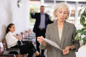 Upset old woman holding white paper, standing in waiting room with her back to person who is...