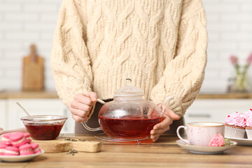 Young woman holding teapot with hibiscus tea in kitchen