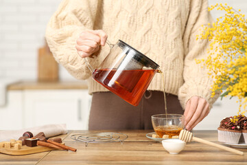 Young woman pouring tea with honey from teapot into cup in kitchen