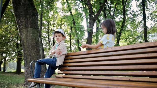 A little preschool boy is being taught not to take candies from strangers. A little boy is sitting on a bench in a park. A girl offers him a treat or candy, but the boy doesn't take it