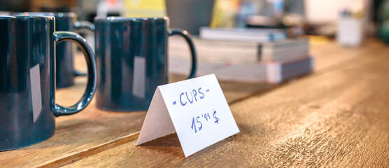 Banner of articles price label over wooden counter in local store with industrial style decoration. Interior of empty vintage shop with breakfast blue mugs on table ready to sell. No People.