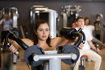 Fototapeta premium Concentrated fit young girl performing set of shoulder exercises in weight machine in modern gym. Physical activity, fitness and sports motivation concept..