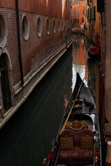 A gondola in Venice, Italy with its ornate wooden interior and black oar,  the narrow canals of an ancient city surrounded by historic buildings with red tiles and white curves on their walls © Rita