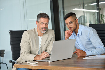 Two busy professional business men working together talking using laptop in office. Male manager and employee looking at computer discussing investment business project analysis at office meeting.