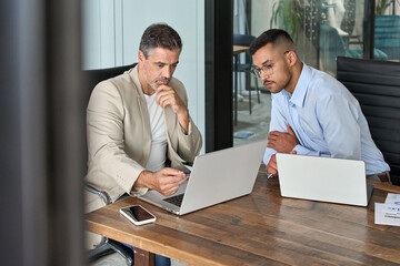 Busy professional team of two business men executive, male partners working using laptop looking at computer talking analyzing online trading financial market data at work in office sitting at desk.