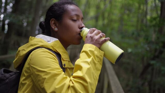 Young Latin woman wearing yellow rain jacket drinking of a sustainable metal water bottle taking a break on a hike on Nature. Active people hiking in the bush and forest. No plastic