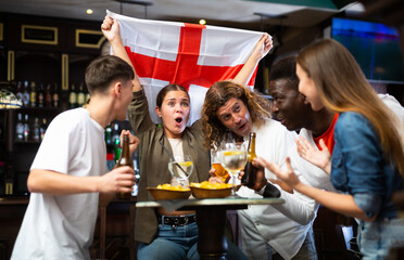 Cheerful multiracial football fans waving the flag of England while drinking beer and watching...