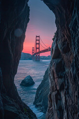 A view of the Golden Gate Bridge from inside, framed by two rocks on both sides, with a pink sky in the background. The bridge is illuminated at dusk and appears red against the blue sea water.  - obrazy, fototapety, plakaty