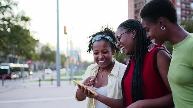 Cheerful African female friends crossing street looking laughing cell phone. Young black women enjoy stroll together in city using mobile on free day outdoor. Positive social relations generation z 