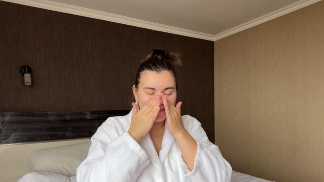 Horizontal 4K video. Young adult woman in white bath robe applies moisturizer to face in hotel room. Concept of rest, skin protection during cold season, cosmetic procedure, spa salon, relax, march 8