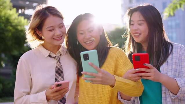 Cheerful Asian female friends walk around city looking laughing cell phone. Young Chinese girls enjoy stroll hugging together using mobile on sunny day outdoor. Positive social relations generation z 