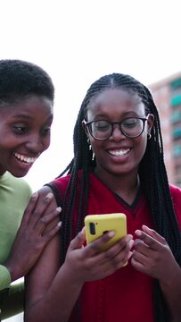 Vertical three nice African American women laughing happy watching funny videos on mobile phone. Young black female friends standing together enjoying free day look cheerful their cell outdoors