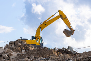A yellow excavator works on top of a pile of construction waste in the process of dismantling a building and structures. A machine with a raised bucket against a sky with light clouds
