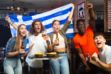 Fototapeta premium Group of cheerful friends sports fans of different nationalities celebrating victory of favorite team in bar, drinking beer and waving national flag of Greece..