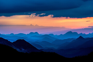 Obraz premium Dark silhouettes of mountains against the sky at sunset with dark clouds. A panoramic view of distant mountain peaks in black silhouettes against the background of a blue and orange twilight sky.