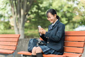 Obraz premium 公園のベンチに座りスマートフォンで動画・SNSを見て笑顔の中学生・高校生の制服姿の女の子 