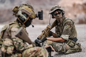 Soldiers in camo gear during a tactical operation with binoculars and rifles on a rocky terrain,...