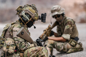 Soldiers in camo gear during a tactical operation with binoculars and rifles on a rocky terrain,...