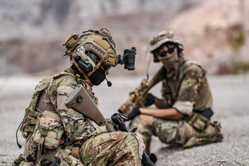 Soldiers in camo gear during a tactical operation with binoculars and rifles on a rocky terrain,...