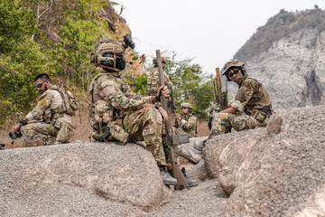 Soldiers in camo gear during a tactical operation with binoculars and rifles on a rocky terrain,...