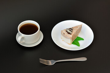 A piece of cream cheesecake with mint on a plate, a cup of hot black coffee and a fork on a black background.