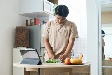 Young man watching online culinary class on tablet computer, making salad with fresh vegetables in the kitchen