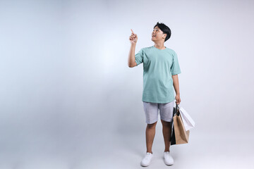 Full length view of young Asian man wear casual summer clothes holding shopping bags and pointing to the empty space for ads