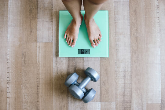 Female feet standing on digital scales for weight control and dumbbells on wooden floor.