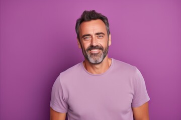 Portrait of handsome mature man in casual t-shirt, looking at camera and smiling while standing against purple background