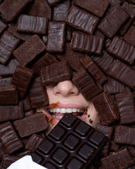The face of a caucasian woman surrounded by chocolates. The girl eats a bar of chocolate.