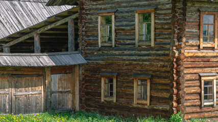 A two-storey old house in the village made of dark logs with a wooden roof. Yellow dandelions in front of the house in the countryside. Wooden old manor house of the 19th century.